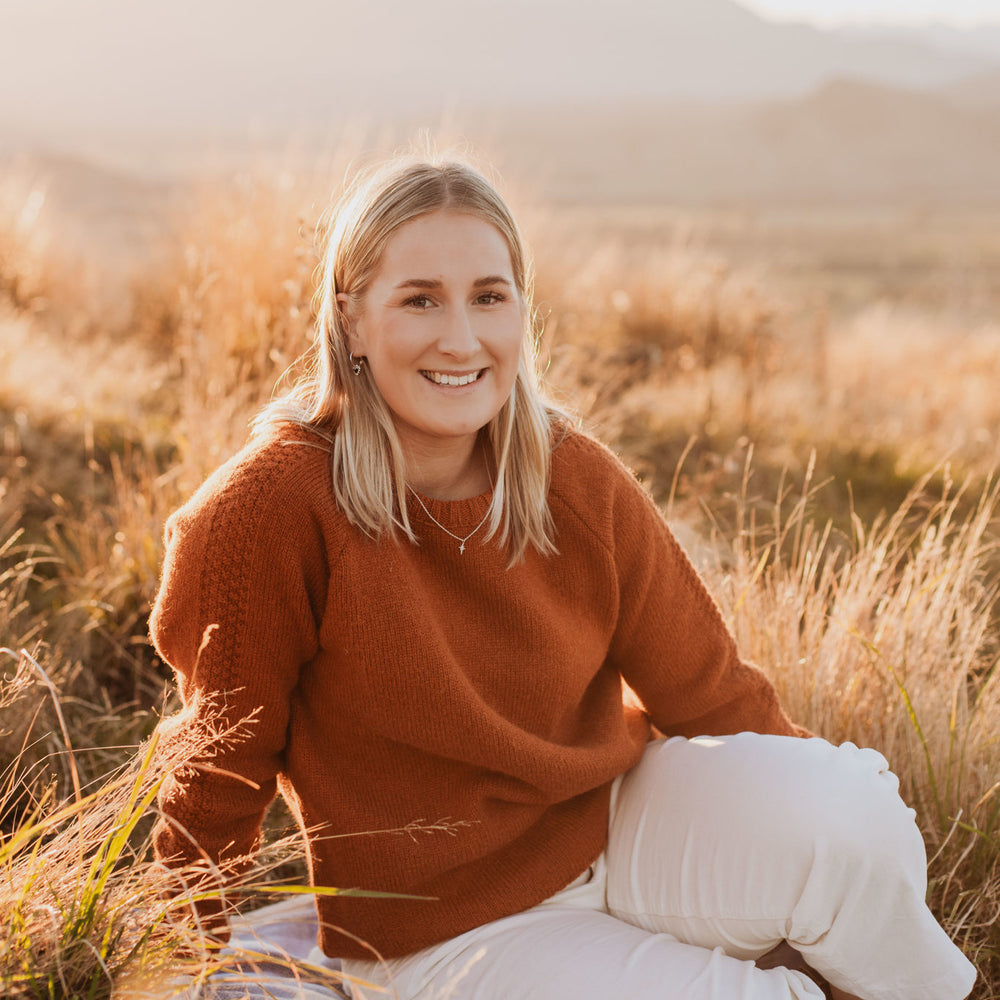 
                      
                        Lady sitting on a wool blanket wearing a McIvor Hill lambs' wool Mabel sweater in rust.
                      
                    