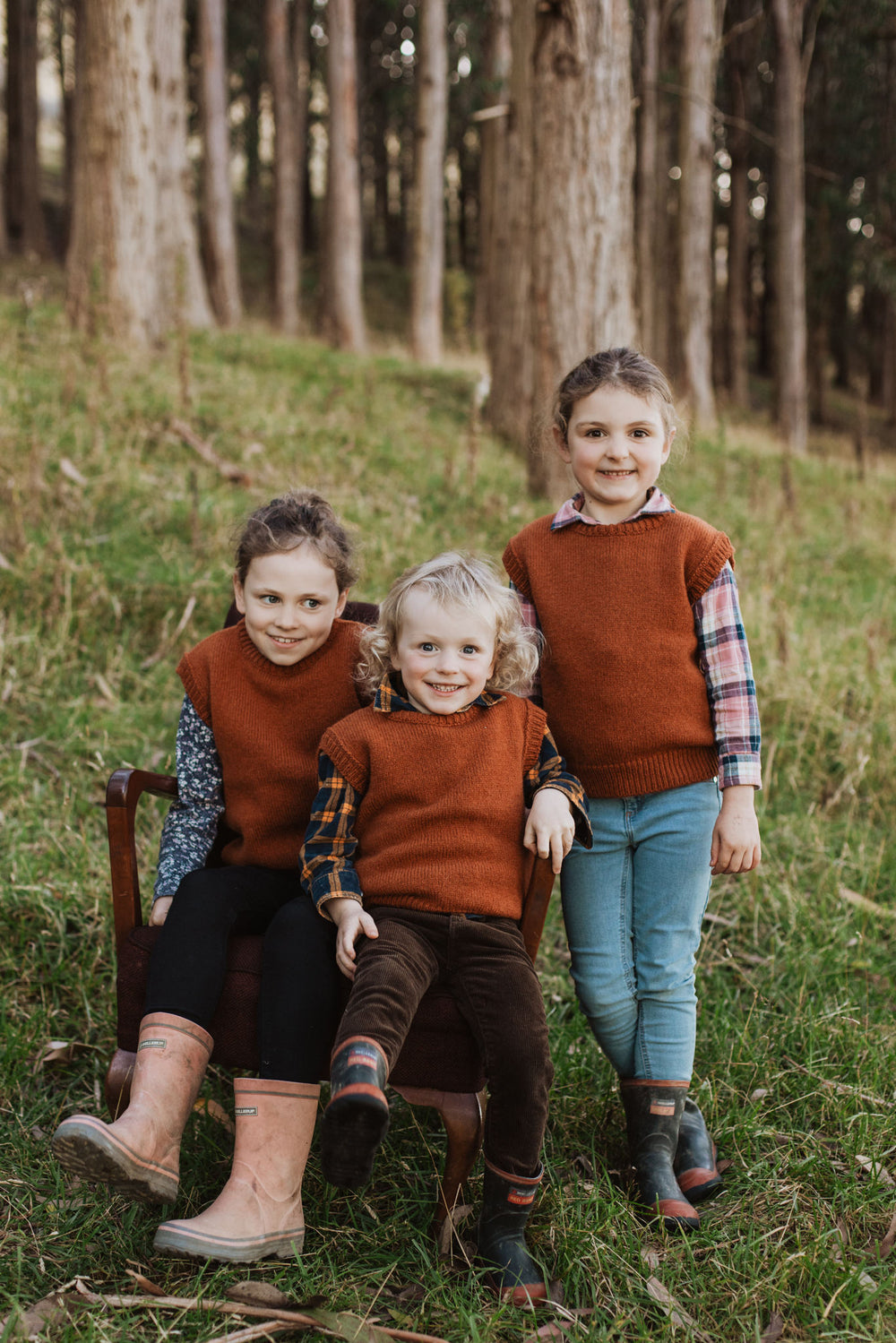 Three children in a forest wearing McIvor Hill lambs' wool knit vests in rust