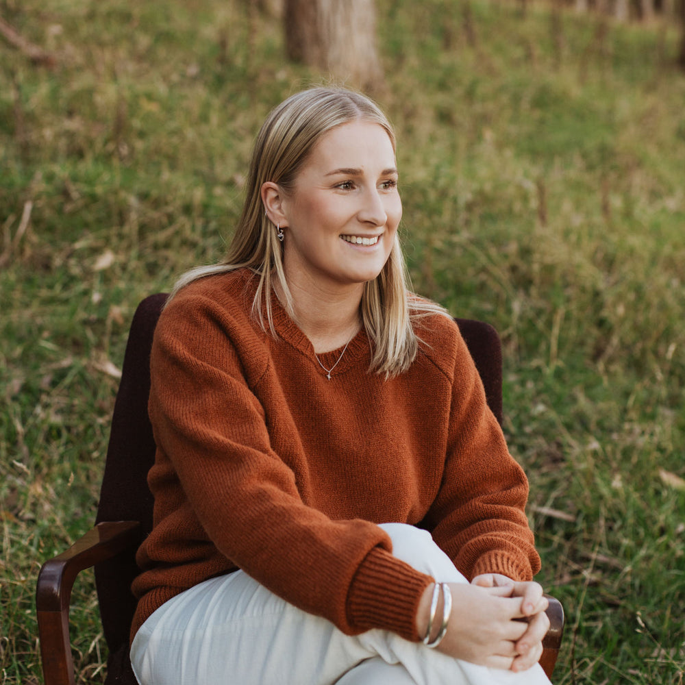 Lady sitting on a chair in the forest wearing a McIvor Hill lambs' wool Rita sweater in rust.