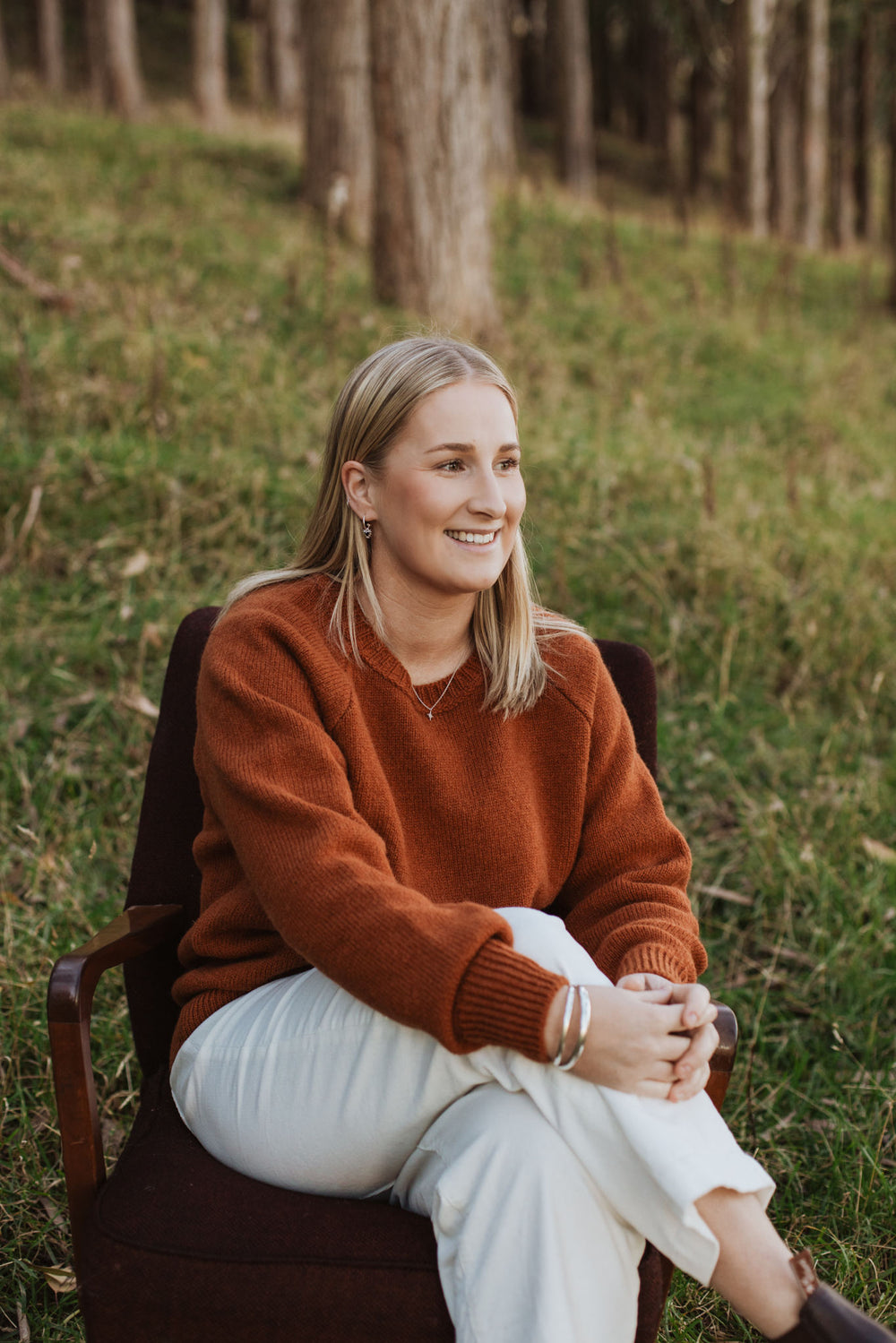 Lady sitting on a chair in the forest wearing a McIvor Hill lambs' wool Rita sweater in rust.