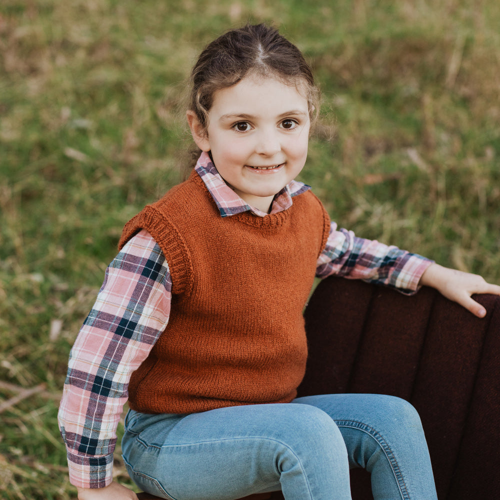 
                      
                        Girl sitting on chair arm in forest wearing a McIvor Hill everyday lambs' wool vest in rust
                      
                    