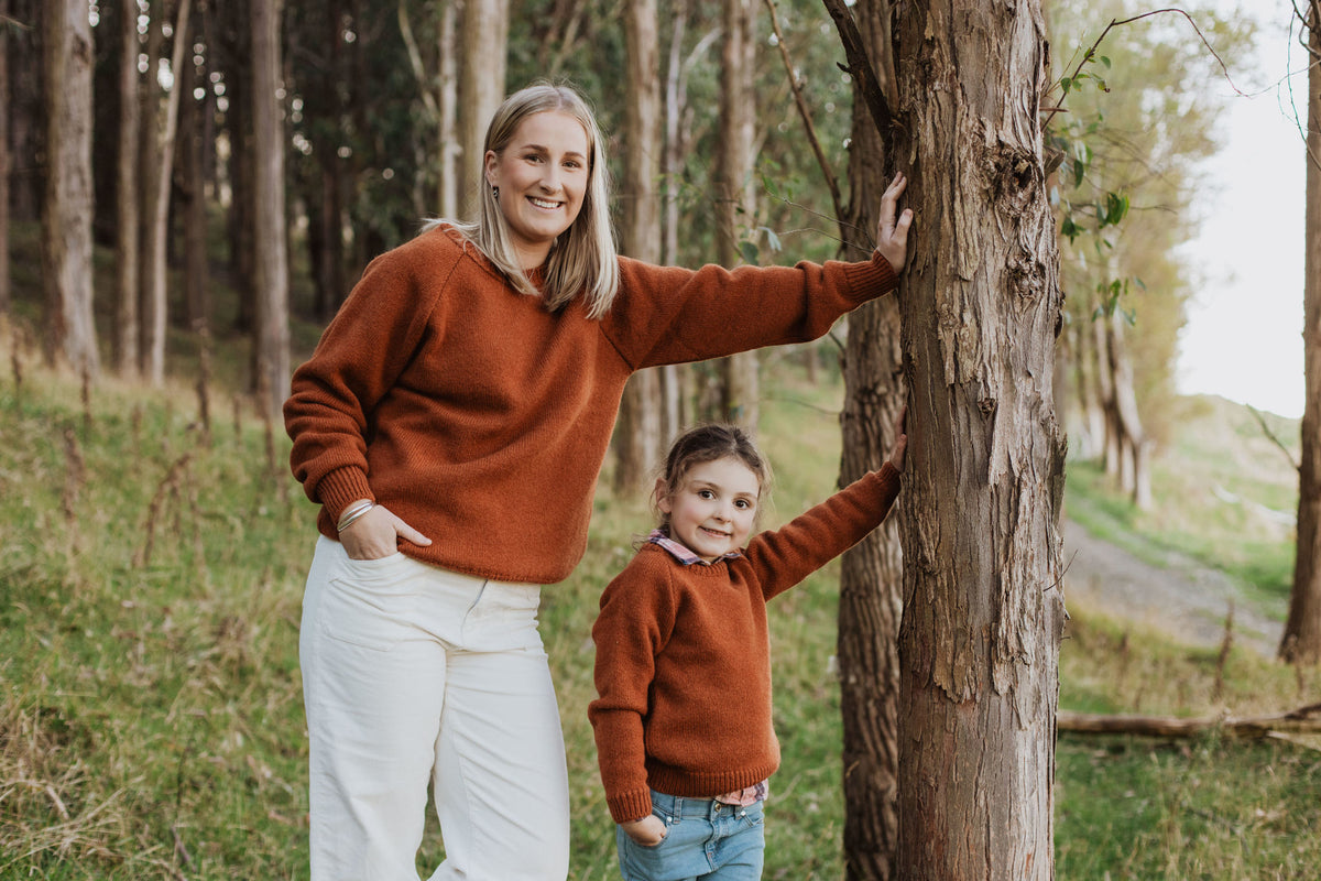 A lady and girl both leaning against a tree wearing McIvor Hill sweaters in rust